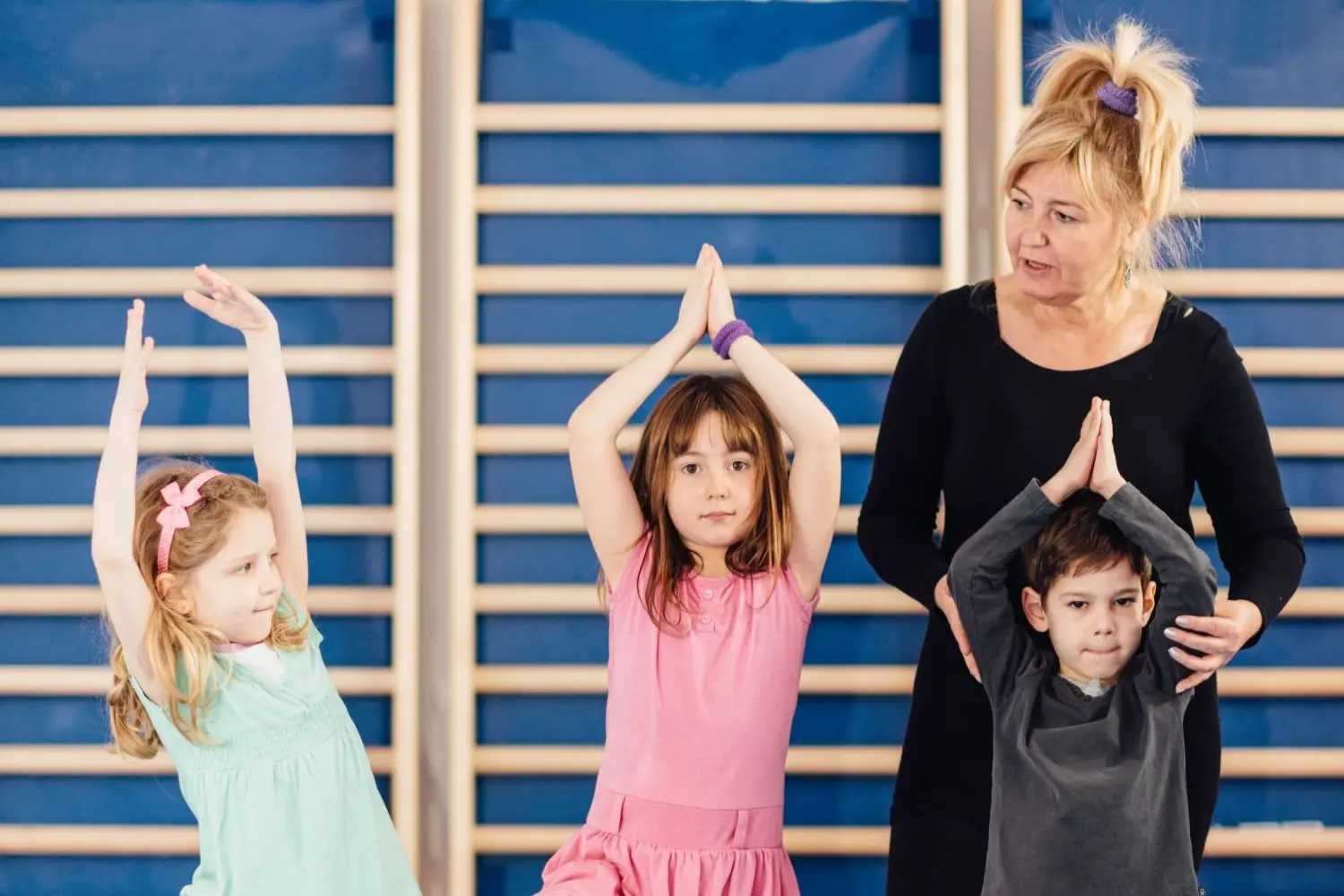 Bewegung, Sport und Mobilität Drei Kinder und eine Lehrerin stehen vor einer Sprossenwand im Turnsaal. Die Kinder halten die Hände gefaltet über den Kopf und nehmen eine Yoga Pose ein