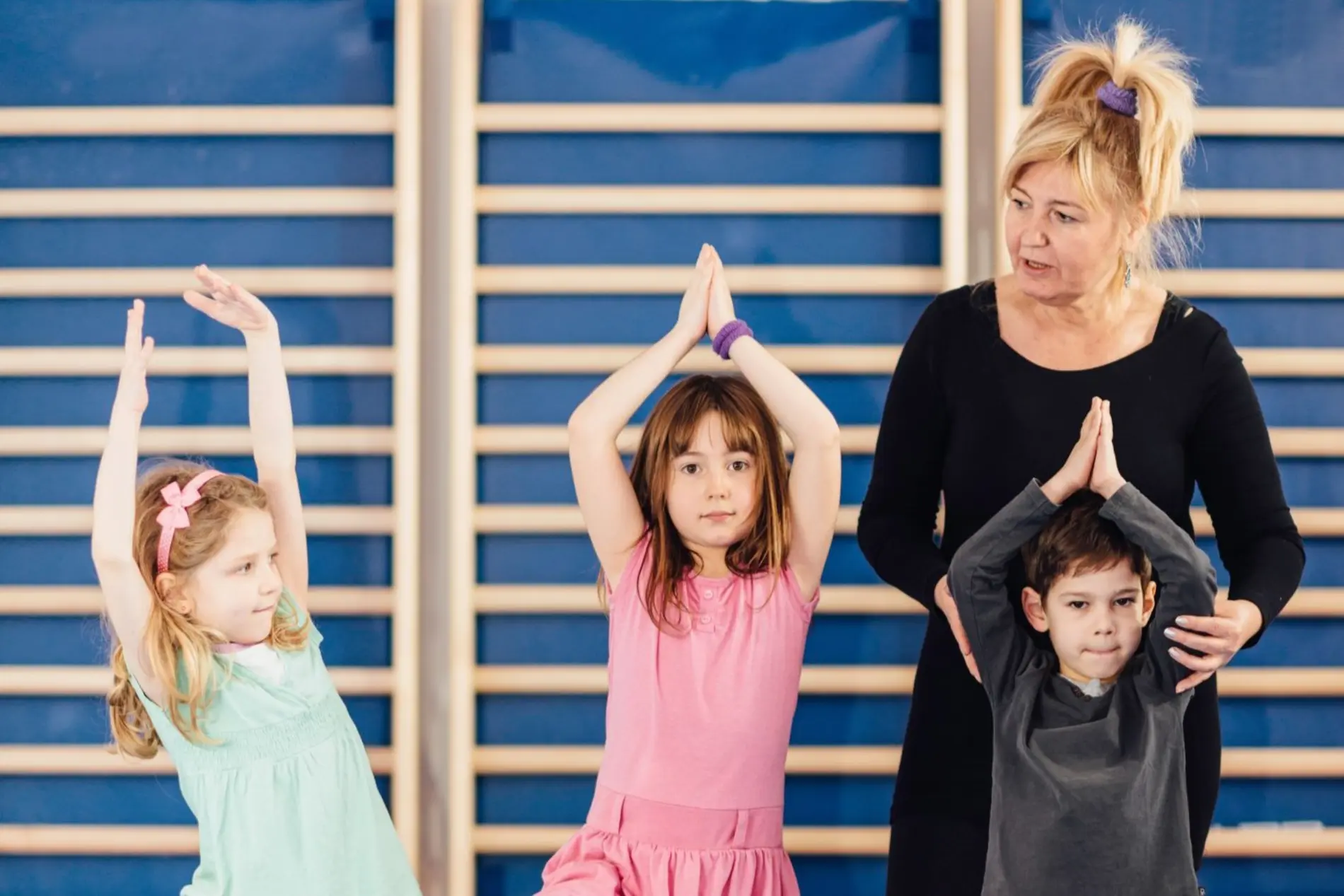 Bewegung, Sport und Mobilität Drei Kinder und eine Lehrerin stehen vor einer Sprossenwand im Turnsaal. Die Kinder halten die Hände gefaltet über den Kopf und nehmen eine Yoga Pose ein