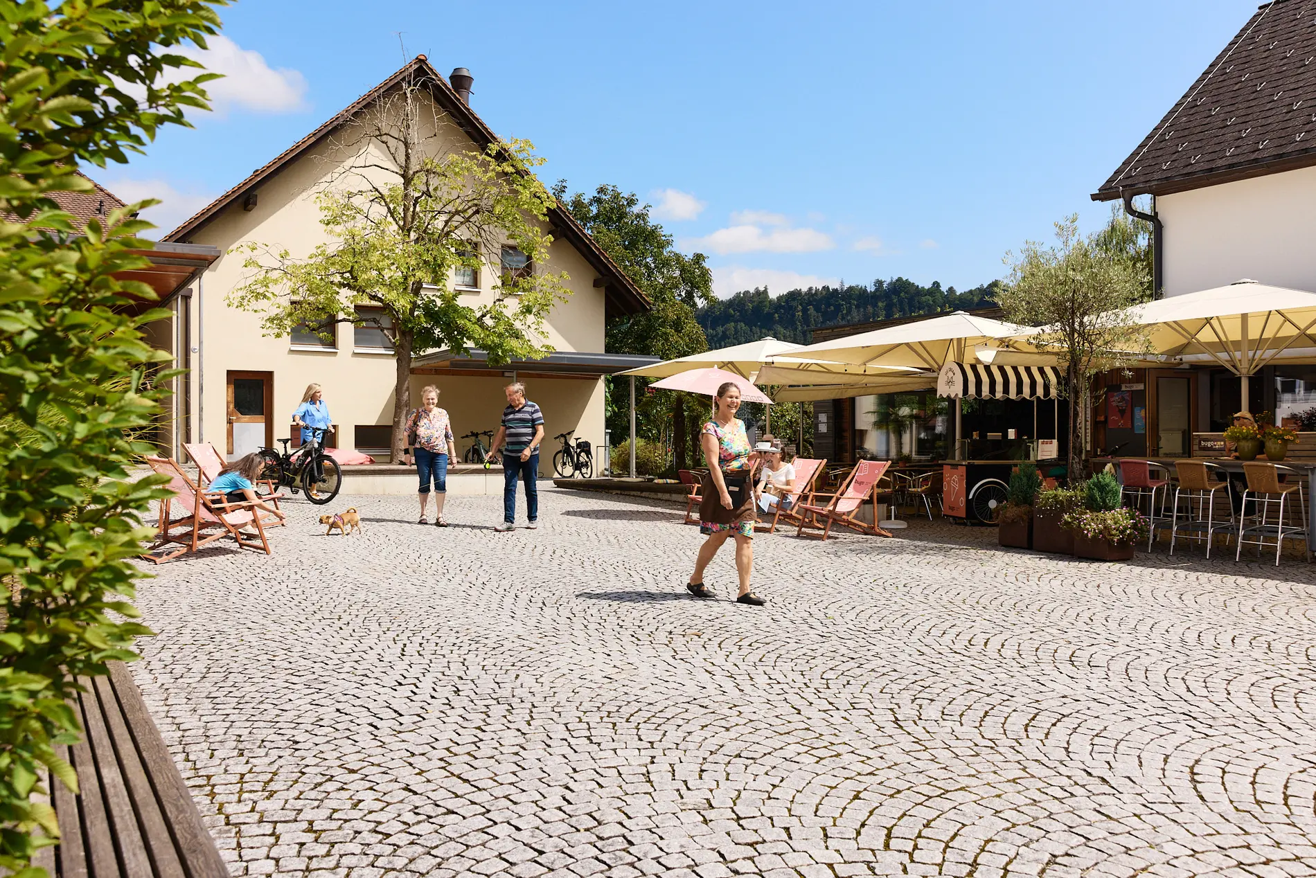 Ein öffentlicher gepflasterter Platz einer kleinen Gemeinde in Österreich. Einzelne Personen gehen vorbei. Rechts ist ein Cafe mit Sitzplätzen im Freien, welche mit Schirmen überdacht sind.