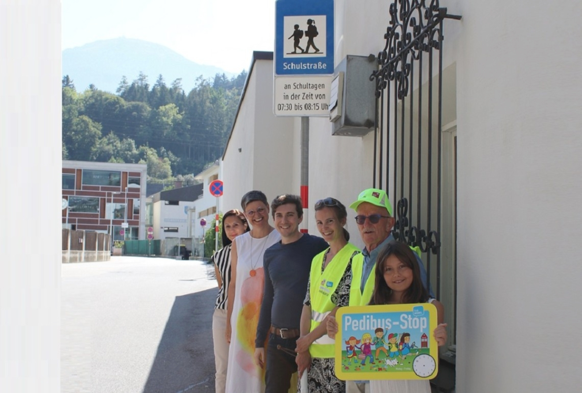 Erwachsene und Kinder stehen mit einem Pedibus-Haltestellenschild direkt vor Beginn des Verkehrsschildes "Schulstraße". Sie lächeln in die Kamera