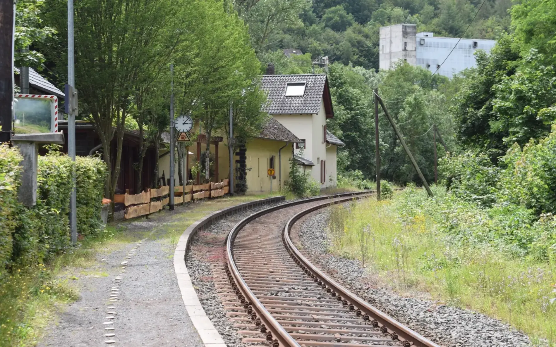 Ein älterer kleiner Bahnhof in der Ferne, vorne der Bahnsteig in einer Kurve. Die Gleise daneben schauen älter aus. Seitlich und im Hintergrund viel Grün