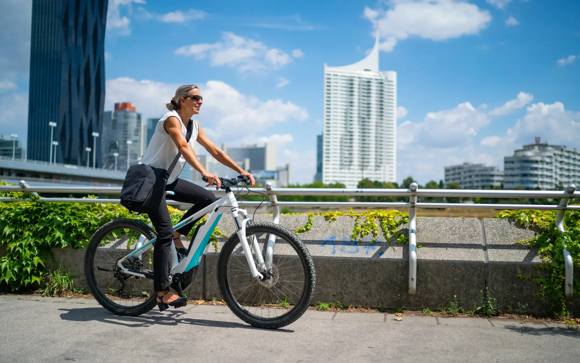 Eine blonde Frau mit Sonnenbrille fährt auf einem E-Bike, im Hintergund sind Hochhäuser und eine Brücke zu sehen.