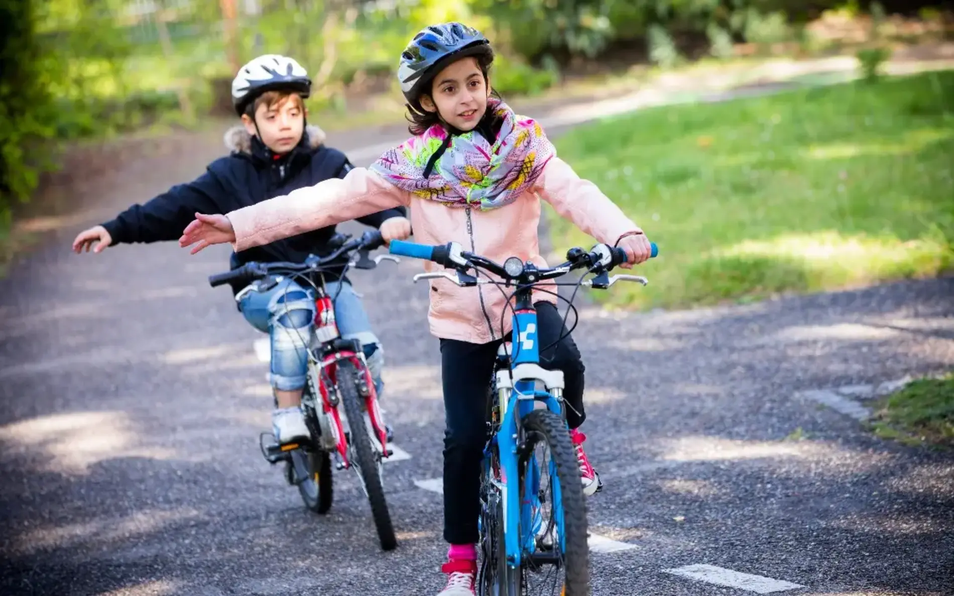 Zwei Kinder fahren Fahrrad und üben das Rechtsabbiegen. Sie halten die rechte Hand seitlich ausgestreckt während sie mit dem Fahrrad fahren. Beide tragen Helm.