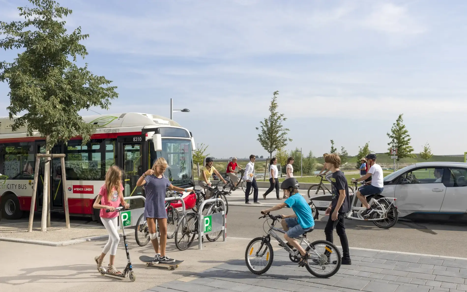 Ein breiter Geh- und Radweg mit Kindern auf Roller und Rad. Dahinter eine Straße mit Bus und E-Auto