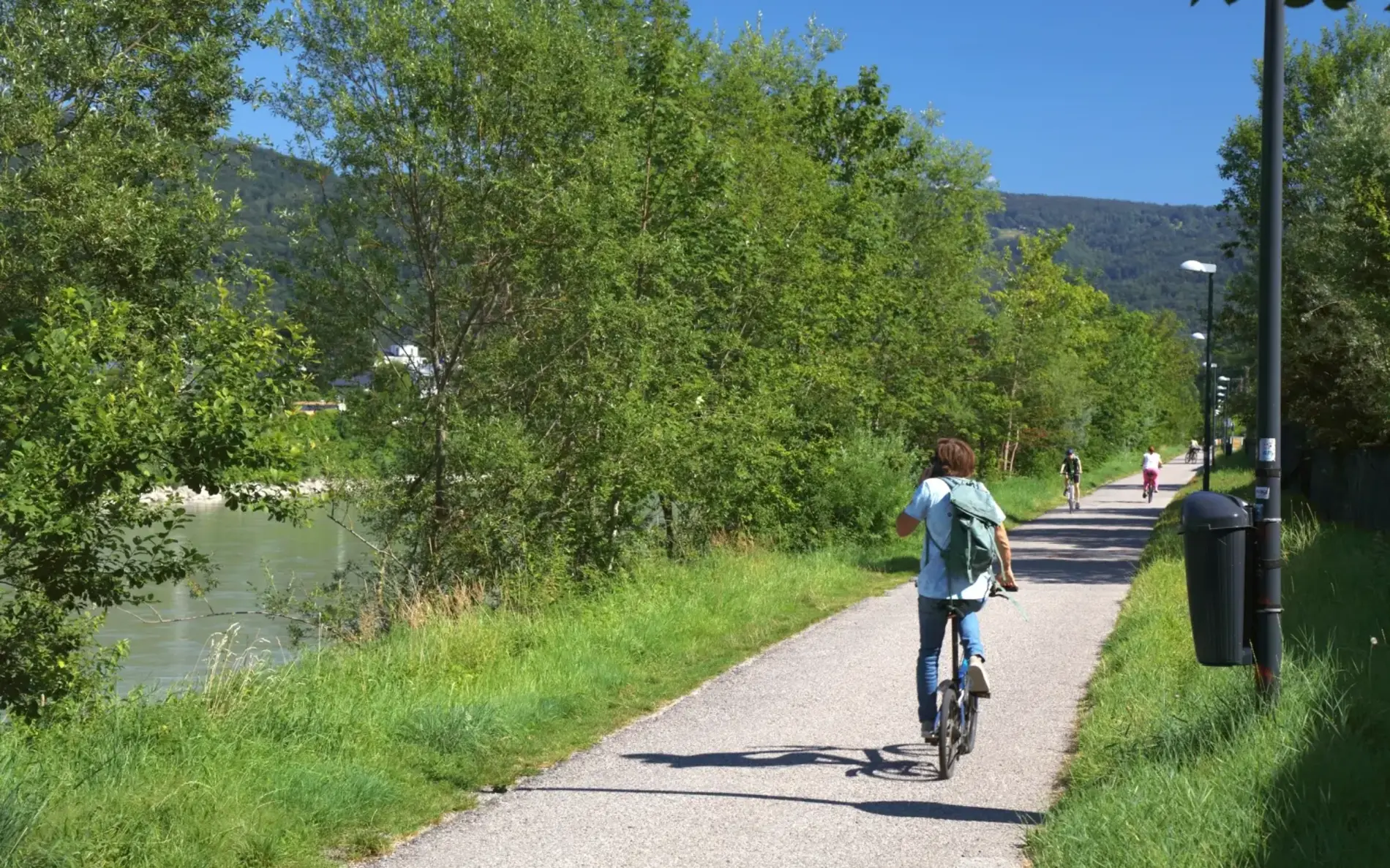 Eine Radfahrerin auf einem Faltrad am Tauernradweg von hinten. Links fließt ein Fluss, direkt neben dem Weg ist alles begrünt und mit Laternen ausgestattet.