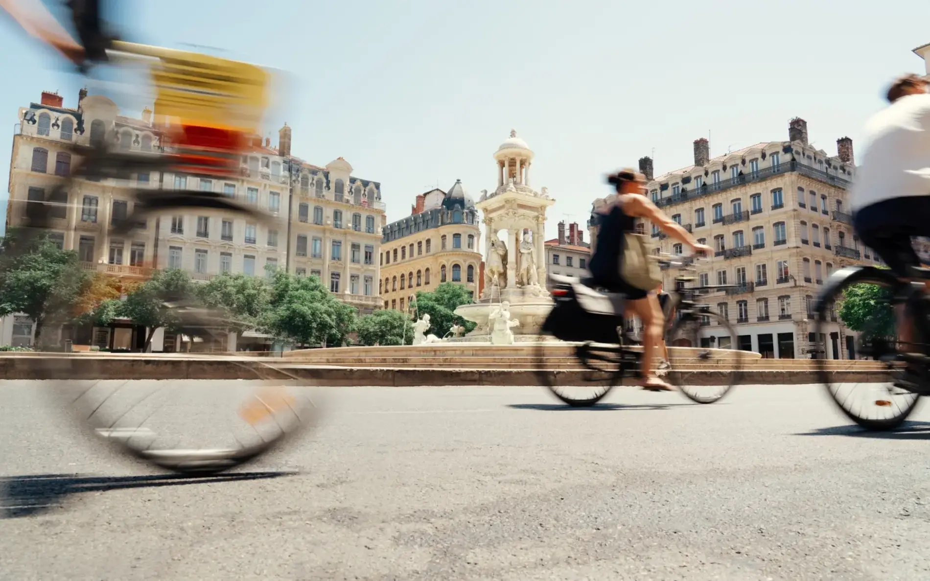 Drei Radfahrende in Bewegung fahren an einem runden Platz in Lyon vorbei. Es scheint die Sonne und die Radfahrenden tragen Sommerkleidung.